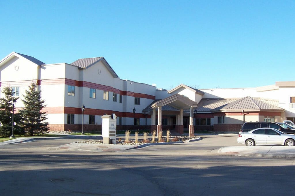 Across the street view of two-story beige and red building with drive up covered entrance and sign in the front that reads "Oak Crossing."