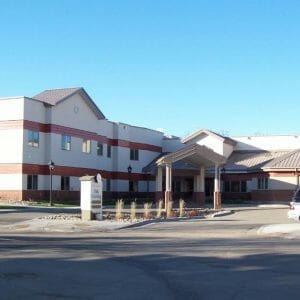 Across the street view of two-story beige and red building with drive up covered entrance and sign in the front that reads "Oak Crossing."