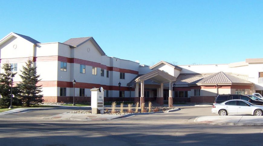 Across the street view of two-story beige and red building with drive up covered entrance and sign in the front that reads "Oak Crossing."