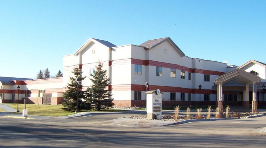 Across the street view of two-story beige and red building with drive up covered entrance and sign in the front that reads "Oak Crossing."