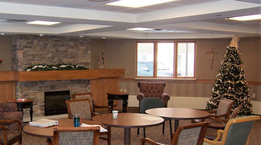 Gathering room with round tables and chairs sitting in front of stone fireplace with lit Christmas tree to the right.