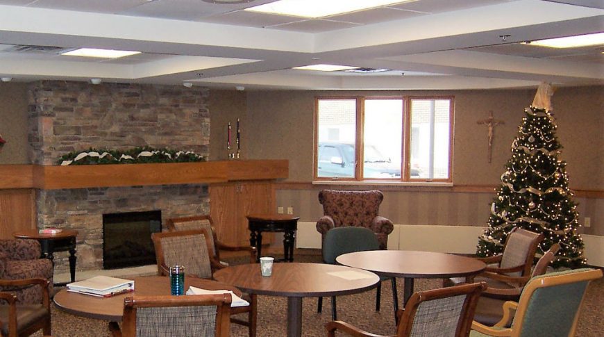 Gathering room with round tables and chairs sitting in front of stone fireplace with lit Christmas tree to the right.