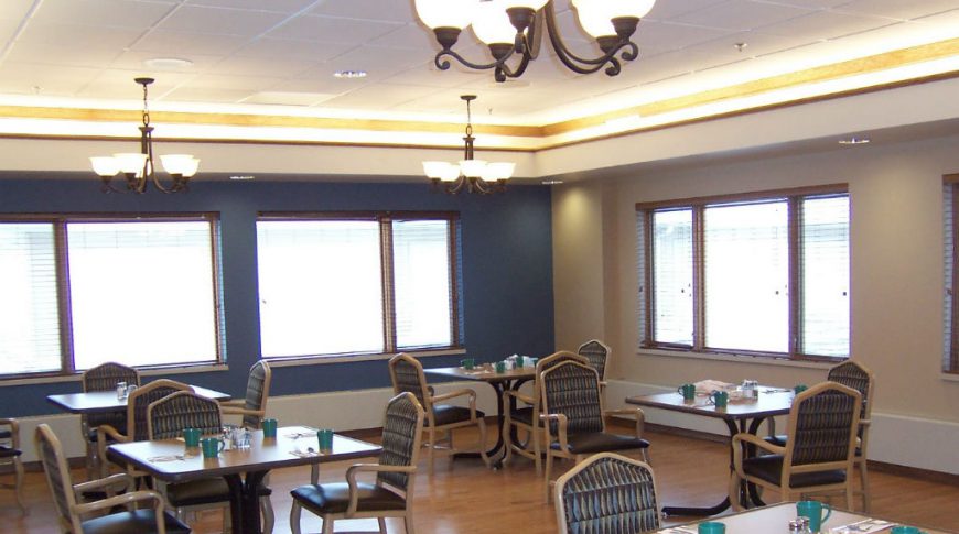 Dining room with square tables and chairs set with silverware and coffee mugs with beige and dark navy accent walls and chandeliers overhead.