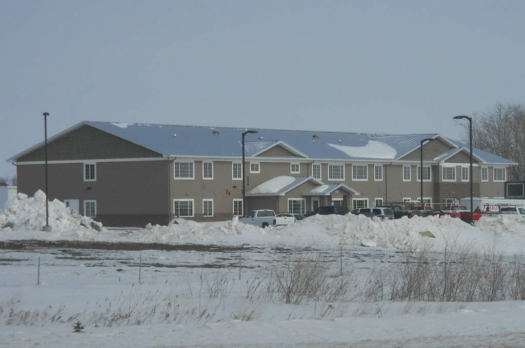 Exterior view from across the street of long rectangular light taupe building with windows throughout, metal roof and awning entrance with parking lot and street lights in front.