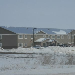 Exterior view from across the street of long rectangular light taupe building with windows throughout, metal roof and awning entrance with parking lot and street lights in front.