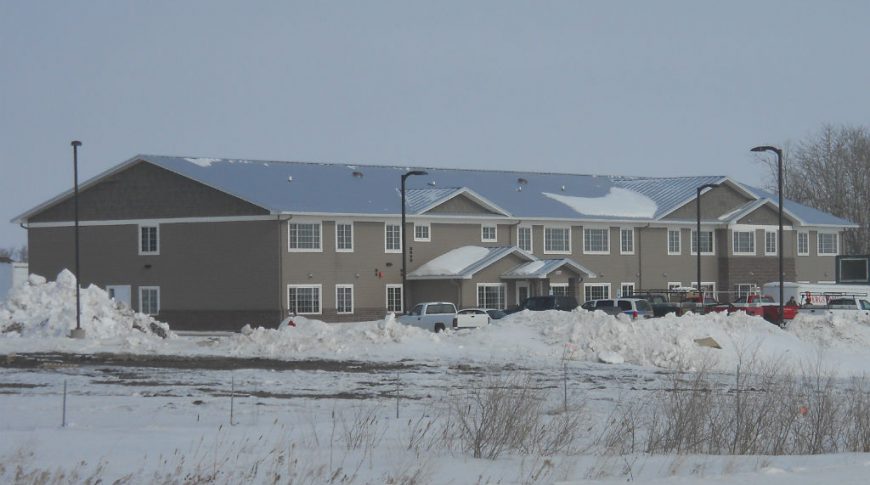 Exterior view from across the street of long rectangular light taupe building with windows throughout, metal roof and awning entrance with parking lot and street lights in front.