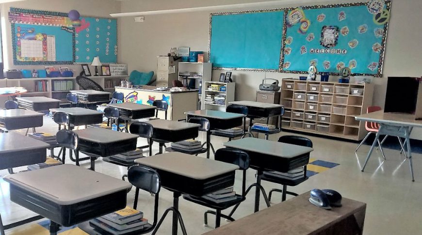 Tioga Elementary School-Int1-1024x680-Education Construction Beige wall classroom with beige floor tiles with blue and yellow accent tiles and individual desk stations in middle of room with cubbies and blue tack boards on outer walls.