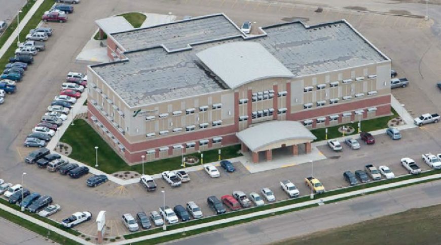 Trinity Health Western Dakota-Ext4-edit-1024x680-Healthcare Construction Overhead aerial view of concrete and red-bricked three-story building with flat roof and large concrete-awning entrance with parking lot surrounding each side.