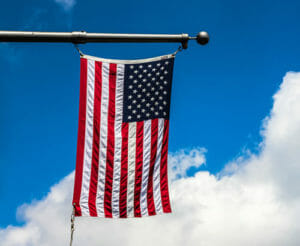 American flag hanging down from metal flagpole with bright blue sky with clouds in the background.