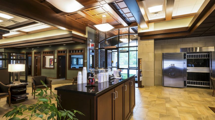 Reception area with light beige tiles underneath, dark wooden cabinetry with coffee station in center, and overhead dark wooden millwork.