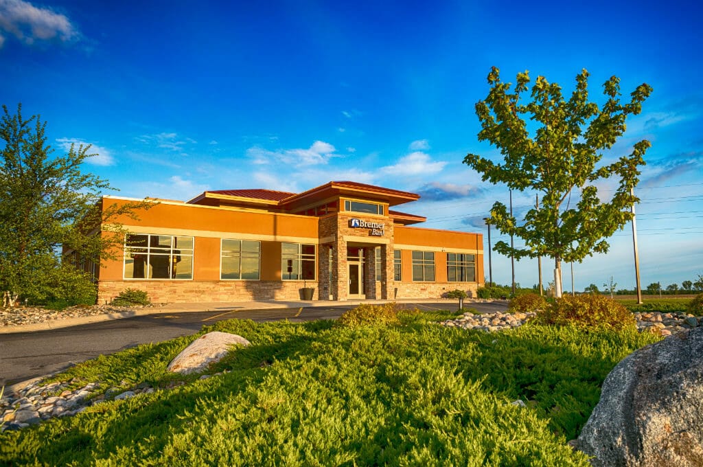 Exterior view of Brown and brick building with awning entrance and green shrubbery, rock, and trees in foreground.