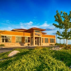 Exterior view of Brown and brick building with awning entrance and green shrubbery, rock, and trees in foreground.