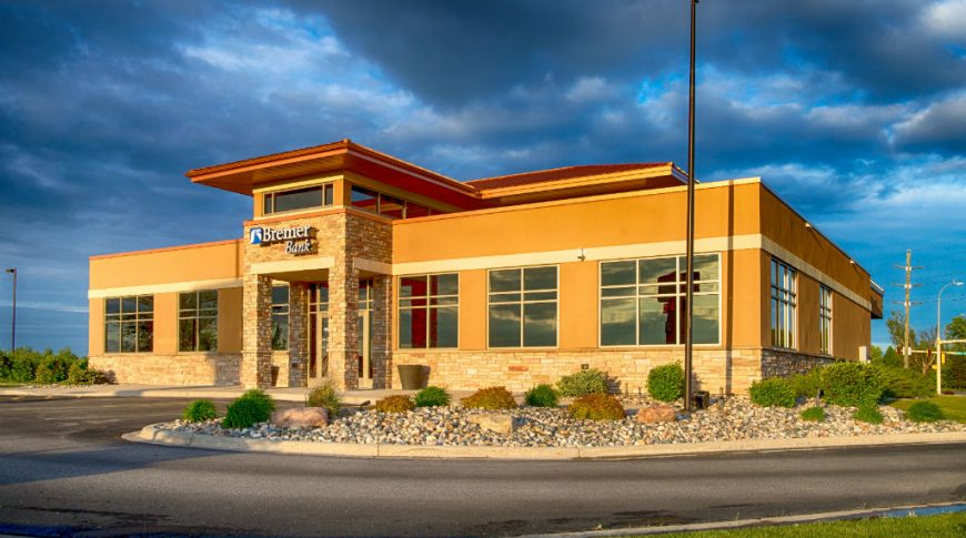 Entrance of brown and beige bricked-building with rock and shrubbery landscaping to exterior.