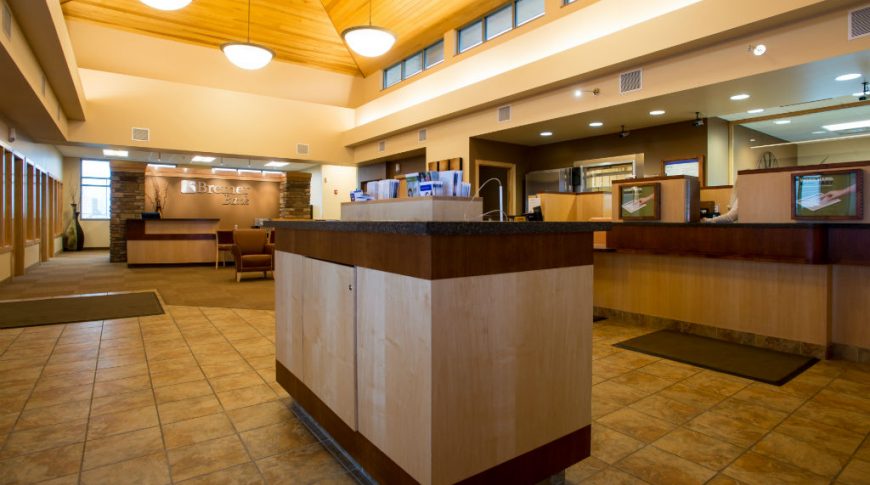 Beige two-story atrium interior of Bremer Bank with light wooden reception desks and black countertops with shades of brown tiled flooring with light oak paneled ceiling above.