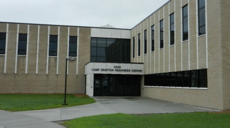 Entryway to two-story light brick building with windows on second floor, floor to ceiling windows in the entryway and windows on first and second floor of building on the right side.