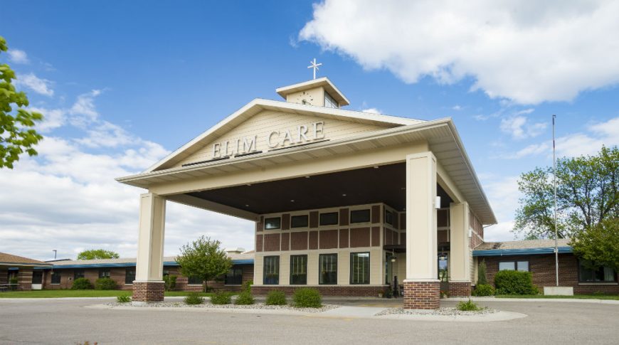 Fargo Elim Health Care Center-P-Ext1-1024x680-Housing Commercial Construction Exterior view of front drive-up entrance to yellow and brick building with awning overhead and the words "Elim Care" above.