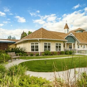 Exterior garden view of Victorian-cottage-looking yellow and green building with light brown shingles, picket fence with lush green grass and flowers and shrubbery surrounding.