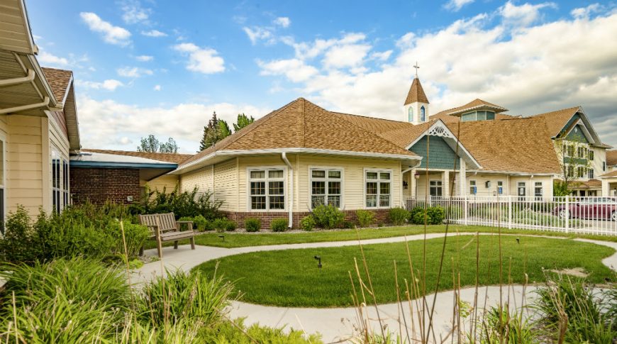 Fargo Elim Health Care Center-P-Ext2-1024x680-Housing Commercial Construction Exterior garden view of Victorian-cottage-looking yellow and green building with light brown shingles, picket fence with lush green grass and flowers and shrubbery surrounding.