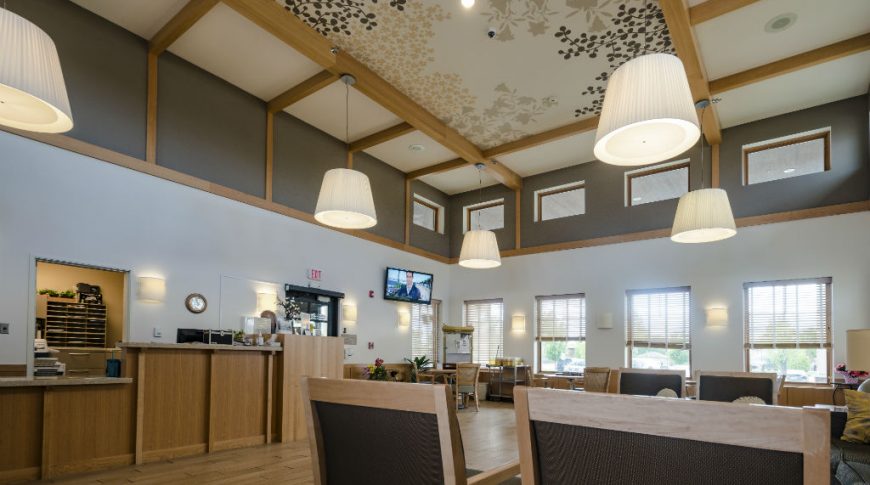 Fargo Elim Health Care Center-P-Int1-1024x680-Housing Commercial Construction Large rectangular 2-story room with upper and lower windows to the back wall, light flooring nd wooden accents throughout, reception area to left and seating chairs and couches to the foreground.