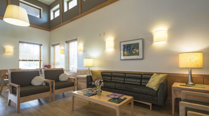 Fargo Elim Health Care Center-P-Int2-1024x680-Housing Commercial Construction Light beige 2-story wall interior with light faux wood plank floors and couches and chairs seating area, Two-Story open area with windows overhead.