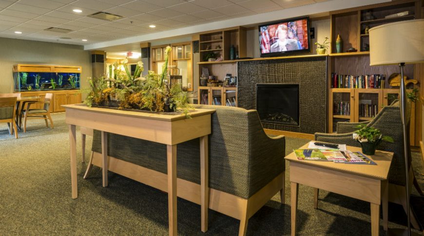 Fargo Elim Health Care Center-P-Int3-1024x680-Housing Commercial Construction Living room interior at assisted living home with fireplace and tv overhead to the back wall, fish tank to back left wall, and sitting couches and chairs in foreground.