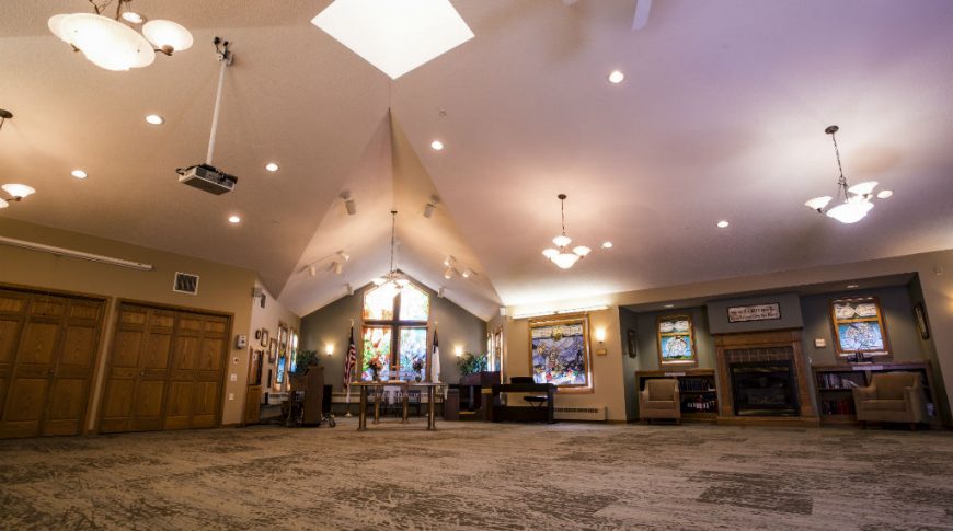 Fargo Elim Health Care Center-P-Int4-1024x680-Housing Commercial Construction Interior chapel with stained glass windows to back center wall and fireplace seating area to back right wall with wooden closet doors to back left wall.