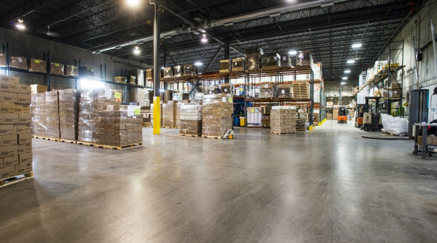 Concrete-floor warehouse with black metal overhead roof and beams and rows of pallets with boxes throughout.