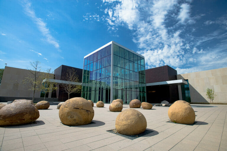 Exterior of modern building with two-story glass atrium and dark brown brick building exterior behind with light beige concrete paneled building exterior behind that with brick paver and stone patio to the front.