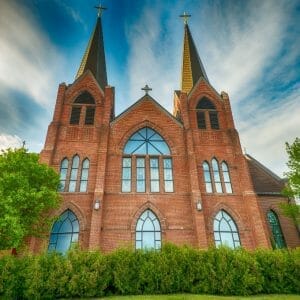Exterior view of large 2-steepled red brick church with large stained glass windows throughout and shrubbery in foreground.