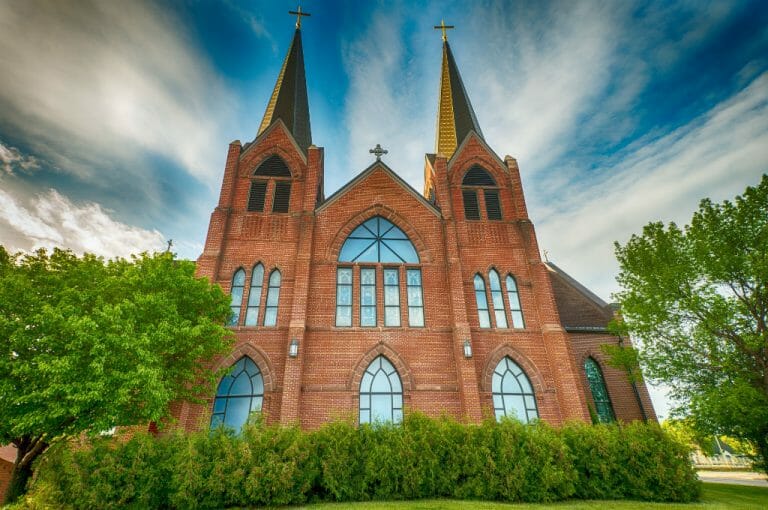Exterior view of large 2-steepled red brick church with large stained glass windows throughout and shrubbery in foreground.