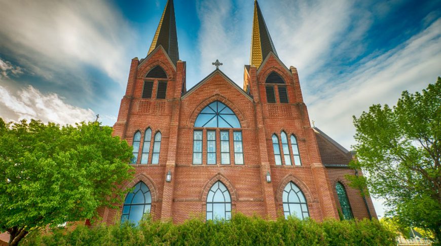 Exterior view of large 2-steepled red brick church with large stained glass windows throughout and shrubbery in foreground.