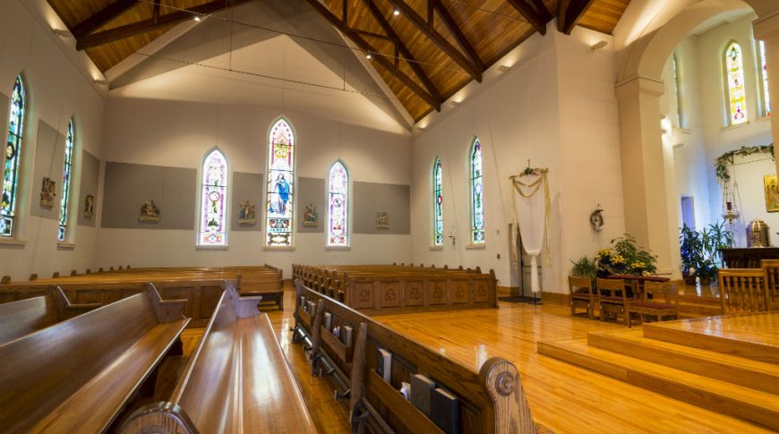 Wooden church pews with large stained-glass windows on each wall with wooden trussed ceiling overhead.