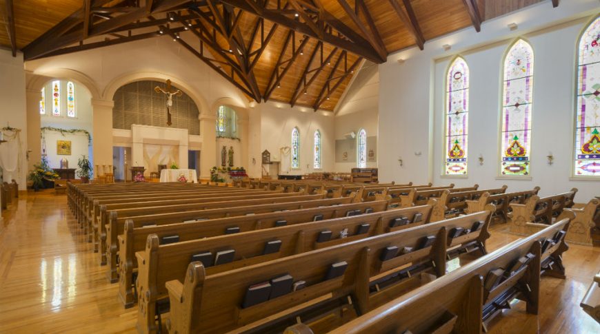 Back corner view of catholic church with light wooden floors and rows of pews, pulpit at the back with large 2-story stained glass windows to the right wall and wooden-planked ceiling and trusses overhead.