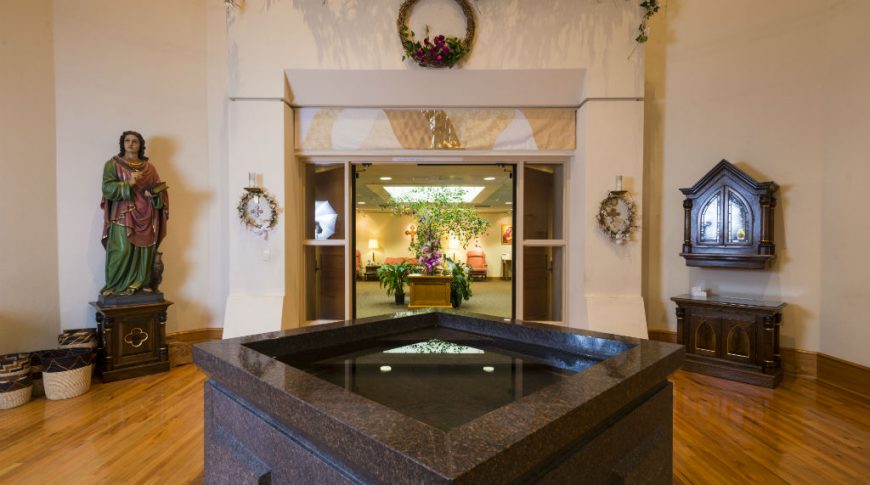 Square granite holy water fountain with statue of saint to back left and cabinetry to back right with church entryway through doors straight back.