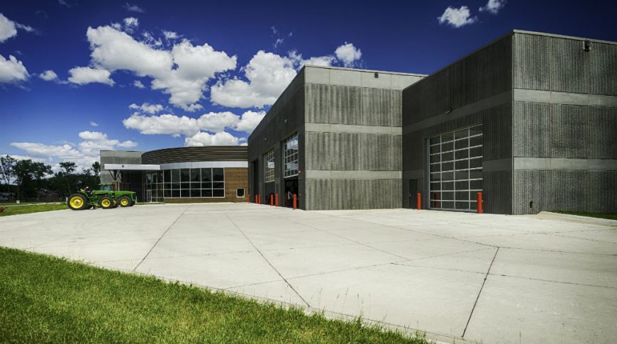 NDSCS Bisek Hall-P-Ext2-1024x680-Education Construction Side view of dark gray precast building with four large equipment full-window garage doors and brown brick curved entryway with floor-to-ceiling windows behind.