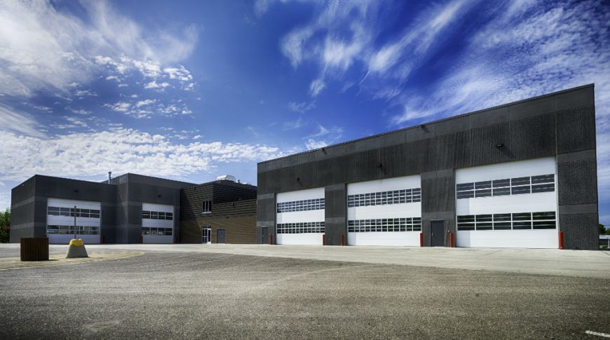 NDSCS Bisek Hall-P-Ext3-1024x680-Education Construction Large equipment garage bays with five large white garage doors with two rows each of windows on dark gray precast concrete building and paved parking lot in foreground.