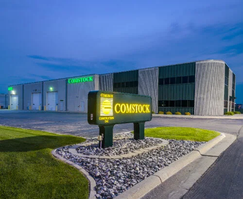 Comstock Construction Fargo-P-500x410-Commercial Construction Fargo, ND's Comstock Construction precast concrete building with lit-up Comstock sign in the foreground at the entrance to the parking lot with blue dusk sky in background.