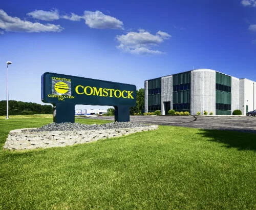 Comstock Construction Fergus Falls, MN-P-500x410--Commercial Construction Comstock Construction precast concrete building in Fergus Falls, MN with dark green panels with Comstock sign in the foreground and bright blue sky with clouds in the background.