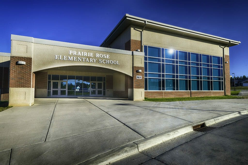 Entrance to school building with the words "Prairie Rose Elementary School" in gold on concrete awning overhead and wall of large windows to the right.