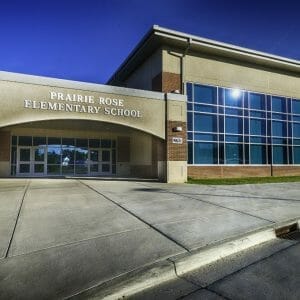 Entrance to school building with the words "Prairie Rose Elementary School" in gold on concrete awning overhead and wall of large windows to the right.