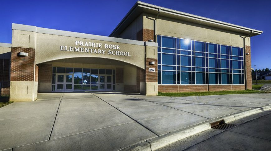 Entrance to school building with the words "Prairie Rose Elementary School" in gold on concrete awning overhead and wall of large windows to the right.