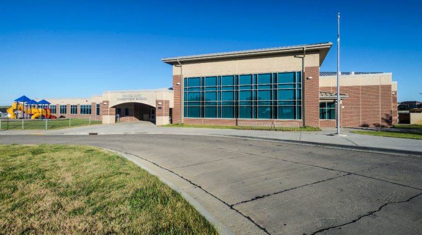 Front drop-off line view of red-bricked elementary school entrance with bright primary-colored playground to back left.