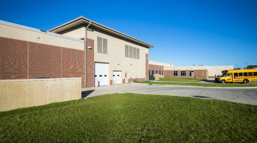 Exterior view of red-bricked school building with two loading bays and small yellow school bus in background.