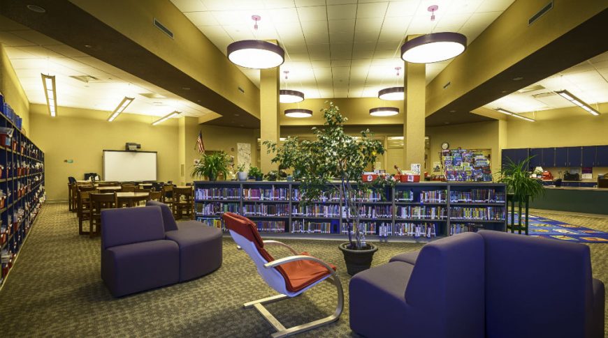 Back corner view of library with large round lights overhead, purple and orange seating in foreground and rows of bookcases to background.
