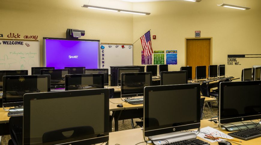 Beige schoolroom with schoolroom-style long narrow desks with computer stations at each chair and a smart board to the back wall.