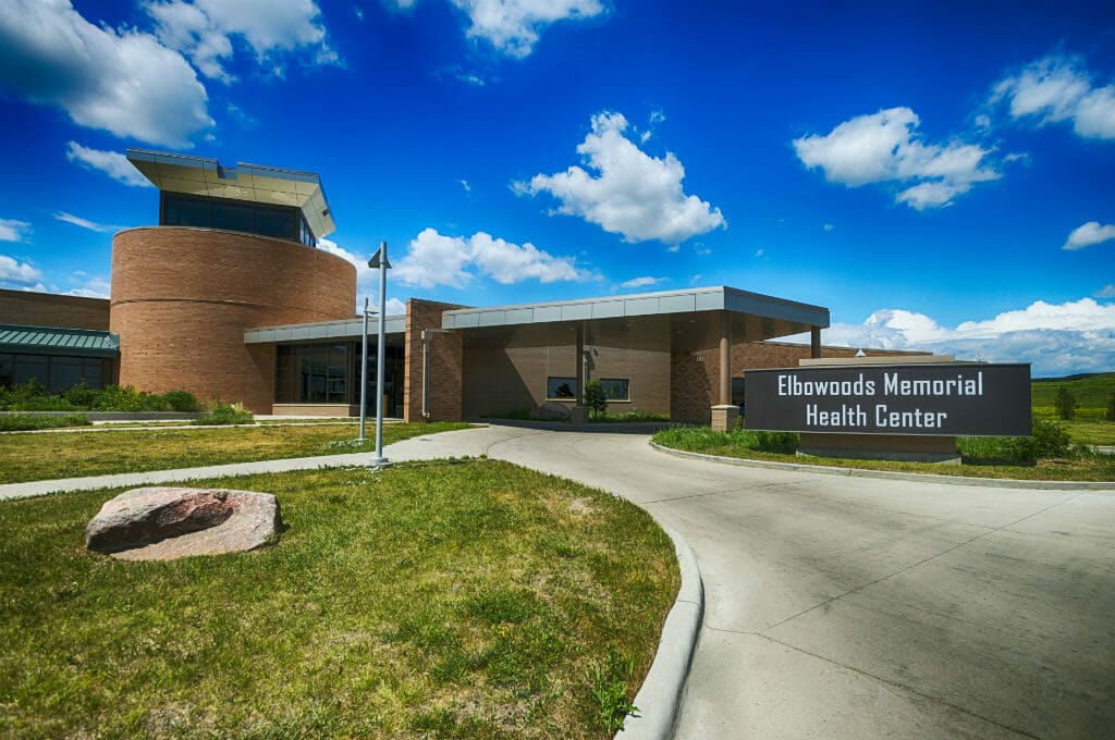 Covered drive-up entrance to brown brick building with protruding second-story rotunda in the back. Sign that reads "Elbowoods Memorial Health Center" to the front right of the building.