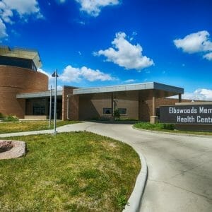 Covered drive-up entrance to brown brick building with protruding second-story rotunda in the back. Sign that reads "Elbowoods Memorial Health Center" to the front right of the building.