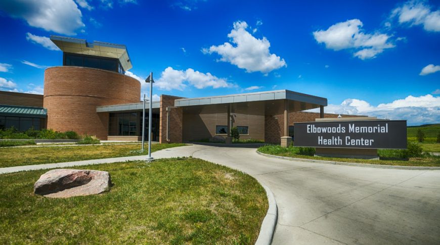 Covered drive-up entrance to brown brick building with protruding second-story rotunda in the back. Sign that reads "Elbowoods Memorial Health Center" to the front right of the building.