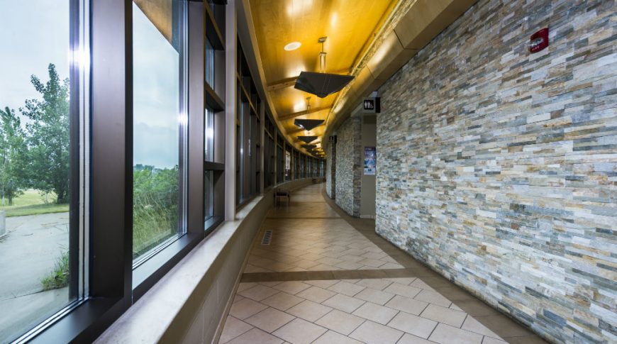 Curved hallway with light slate brick wall on the right and wall of windows to the left overlooking some green trees, shrubs, and gras. Below is a beige and brown tile floor laid diagonally and overhead is a wooden ceiling with chandelier lights up lighting it.