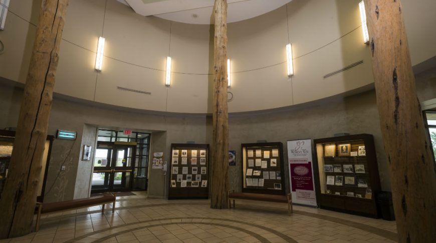 Round rotunda two-stories tall with three wooden posts holding it up, tile floor below with circular pattern in it, lit showcase cabinets around the rotunda walls and second story of rotunda has tall, long lights all around.